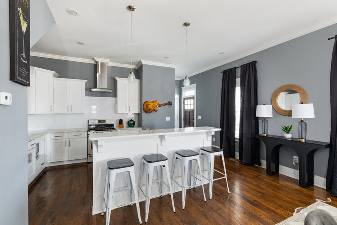 Stylish contemporary home kitchen interior with white cabinets, subway tile backsplash and stainless steel range, marble breakfast bar with four metal stools, gray walls, wood floors, pendant lights, black curtains, console table with lamps and round mirror, and a decorative guitar on the wall.