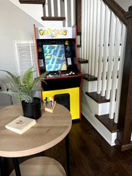 Retro Pac-Man arcade cabinet tucked into a home stairway corner with dark wood steps and hardwood floor, beside a round wooden table holding a potted fern, stacked coasters and colorful golf tees.