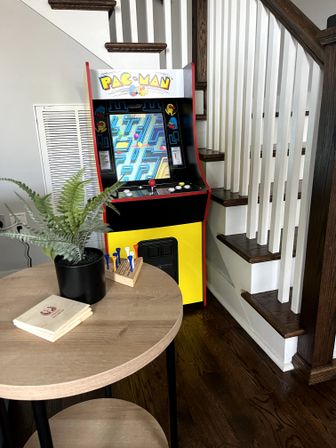 Retro Pac-Man arcade cabinet tucked into a home stairway corner with dark wood steps and hardwood floor, beside a round wooden table holding a potted fern, stacked coasters and colorful golf tees.