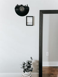Minimalist home interior featuring a half vinyl record cut into a playful city skyline above a small framed print, tall black mirror, and striped vase with eucalyptus on a hardwood floor.