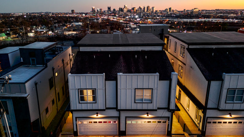 Aerial dusk view of contemporary townhouses with lit double garages in the foreground and a glowing downtown skyline on the horizon, city lights and evening sky visible.