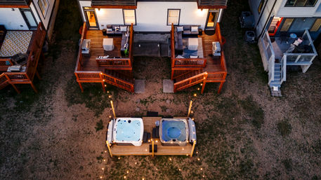 Aerial evening view of two adjacent townhome wooden decks with cozy outdoor seating and twin lit hot tubs on a shared platform, string lights leading across the backyard.