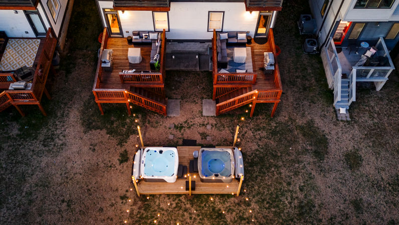 Aerial evening view of two adjacent townhome wooden decks with cozy outdoor seating and twin lit hot tubs on a shared platform, string lights leading across the backyard.