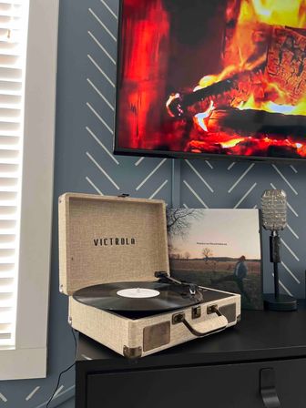 Cozy living room corner with a beige portable record player spinning vinyl on a black dresser, vintage-style microphone and album sleeve behind it, flat-screen TV showing a roaring fireplace, blue patterned wall and window blinds.