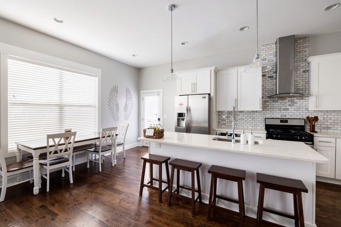Airy modern white kitchen with large quartz island and four dark wood stools, stainless steel refrigerator and range, gray subway tile backsplash, adjacent breakfast nook with white chairs and bench, and warm hardwood floors.