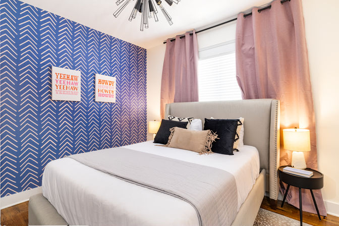 Bright modern bedroom with upholstered gray headboard, white and gray bedding, navy chevron accent wall with playful Yeehaw and Howdy prints, mauve curtains, layered decorative pillows, bedside lamps and black round nightstand.