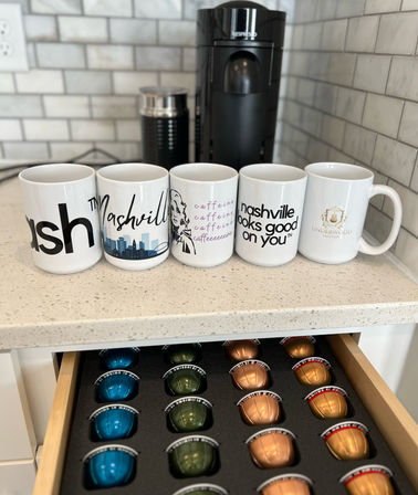 Kitchen coffee station with five Nashville-themed mugs lined up in front of a black pod coffee machine and an open drawer of colorful coffee capsules.