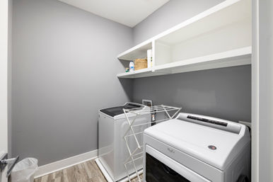 Minimal modern laundry room with white top-load washer and dryer, light gray walls, open white shelves holding detergent and a wicker basket, foldable drying rack and wood-look vinyl floor — bright, tidy, ready-to-fold.