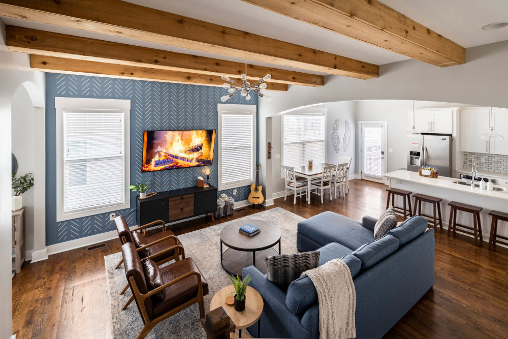 Contemporary farmhouse-style open-concept living room and kitchen with exposed wooden beams, blue accent wall with wall-mounted TV showing a fireplace, hardwood floors, blue sectional sofa, leather armchairs, round coffee table, dining nook, and white kitchen island with bar stools.