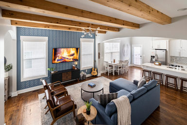 Contemporary farmhouse-style open-concept living room and kitchen with exposed wooden beams, blue accent wall with wall-mounted TV showing a fireplace, hardwood floors, blue sectional sofa, leather armchairs, round coffee table, dining nook, and white kitchen island with bar stools.