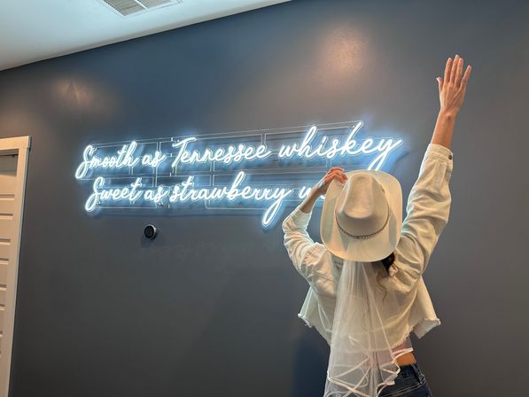 Back view of someone in a white cowboy hat and veil, arms raised toward a bright neon sign on a dark gray wall reading “Smooth as Tennessee whiskey, Sweet as strawberry” — playful country bridal vibe.