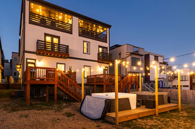 Cozy dusk view of modern coastal-style multi-story vacation homes with illuminated balconies, wooden decks, string lights across the backyard, and two hot tubs on a raised platform.