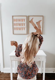 Blonde woman seen from behind sitting at a white desk in a bright modern farmhouse home office, holding a pink mug and wearing a floral dress, with framed wall art above reading “HOWDY” and a cowboy boots print.