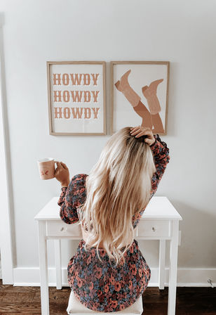 Blonde woman seen from behind sitting at a white desk in a bright modern farmhouse home office, holding a pink mug and wearing a floral dress, with framed wall art above reading “HOWDY” and a cowboy boots print.
