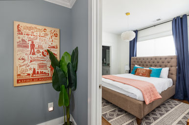 Cozy modern bedroom with tufted beige bed, coral throw and teal accent pillows, navy curtains, patterned rug, tall potted plant, and a vintage-style “Spirit of Nashville” poster on a gray wall.