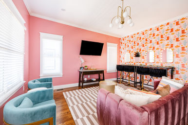 Bright beauty lounge interior with pink walls, playful cowboy-boot and cactus wallpaper, illuminated vanity mirrors, wall-mounted TV, velvet mauve sofa, teal accent chairs and striped rug.