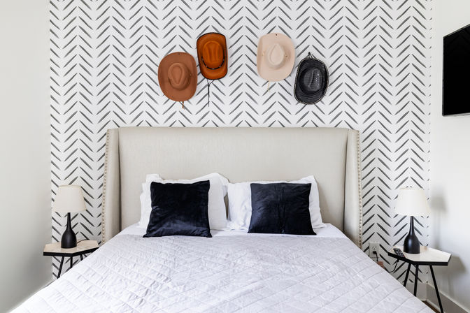 Bright modern bedroom interior with upholstered headboard, chevron-patterned accent wall and four cowboy hats hung above a neatly made bed with white quilt, black throw pillows and matching bedside lamps.