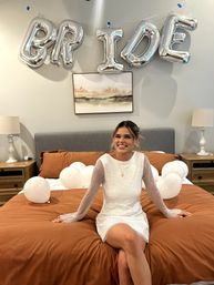 Smiling person in a white dress sits on rust-orange bedding beneath silver "BRIDE" balloons and scattered white balloons in a modern bedroom — bridal celebration.