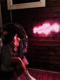 Moody pink-lit scene: person sitting in a hanging wicker chair wearing a cowboy hat, denim fringe jacket, lace top and white boots; neon sign on a wooden wall reads You're like really pretty.
