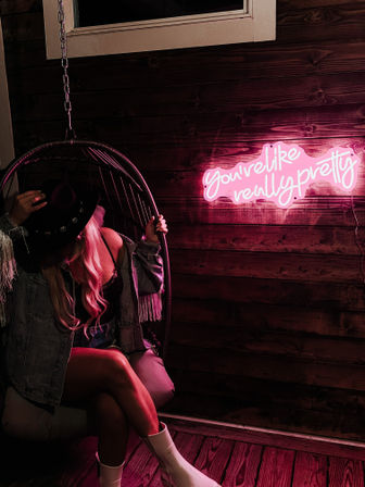 Moody pink-lit scene: person sitting in a hanging wicker chair wearing a cowboy hat, denim fringe jacket, lace top and white boots; neon sign on a wooden wall reads You're like really pretty.