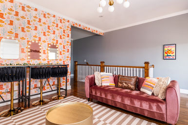 Upstairs loft sitting area with pink velvet sofa, colorful western cactus-and-boot wallpaper, three black velvet bar stools with gold bases at a lighted mirrored bar, striped rug and round brass coffee table.