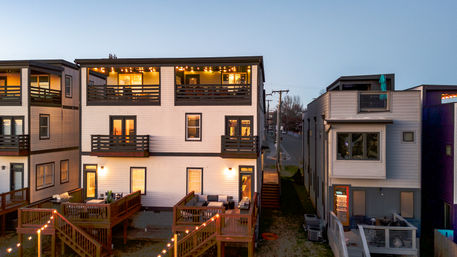Three-story modern townhouses in a residential row with wooden decks, upper balconies, cozy string lights and glowing windows at dusk