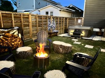 Cozy evening backyard with a glowing fire pit surrounded by Adirondack chairs and tree‑stump seats, stacked firewood, a wooden privacy fence, striped towels, and an illuminated hot tub.