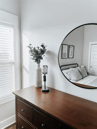 Minimalist bedroom vignette: wooden dresser topped with a white ceramic vase of eucalyptus and a vintage-microphone lamp, round wall mirror reflecting a bed with pillows, sunlight streaming through white blinds.