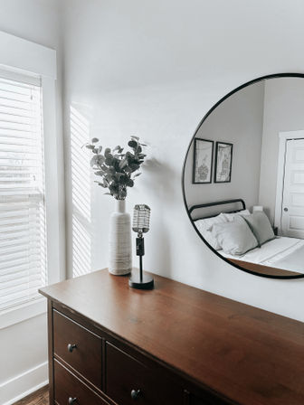 Minimalist bedroom vignette: wooden dresser topped with a white ceramic vase of eucalyptus and a vintage-microphone lamp, round wall mirror reflecting a bed with pillows, sunlight streaming through white blinds.
