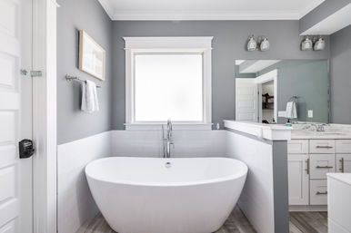 Modern gray master bathroom with a spa-like freestanding white soaking tub under a frosted window, chrome floor faucet, white subway tile and vanity with mirror and wall sconces.