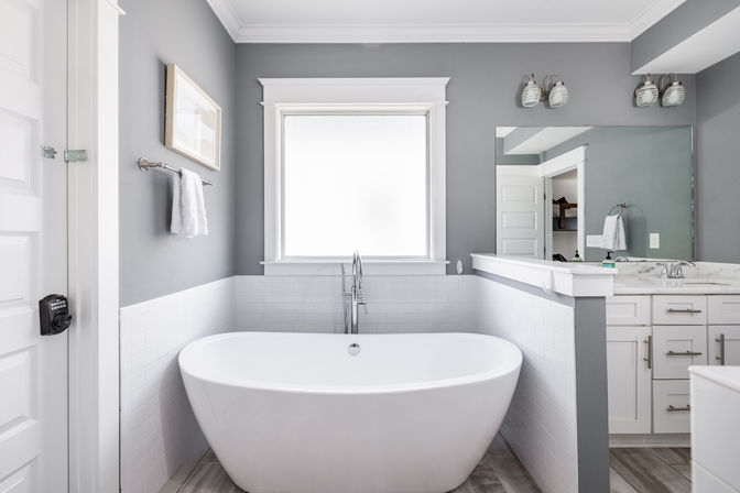 Modern gray master bathroom with a spa-like freestanding white soaking tub under a frosted window, chrome floor faucet, white subway tile and vanity with mirror and wall sconces.