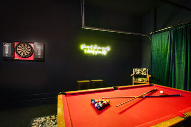 Moody indoor game room: red-felt pool table with racked balls and cues, wall-mounted dartboard, glowing green neon sign, patterned chair and dark green curtains.