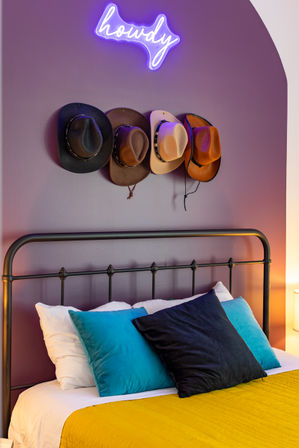 Cozy modern bedroom with black metal bedframe, mustard quilt, teal and navy pillows, purple arched accent wall, a row of cowboy hats on hooks and a glowing neon "howdy" sign above.