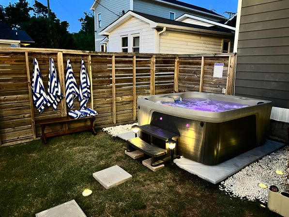 Evening backyard with an illuminated hot tub glowing purple beside a wooden privacy fence, navy-and-white striped towels on a bench, stone pavers and soft landscape lights.