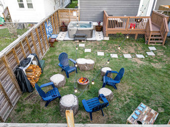 Aerial view of a suburban backyard with a central fire pit encircled by blue Adirondack chairs and tree-stump seats, string lights overhead, a hot tub by a wooden deck, and stacked firewood.