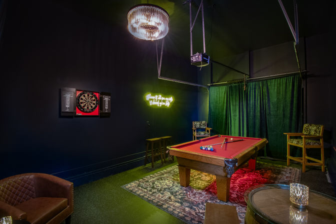 Cozy indoor game room with red-felt pool table and cues, racked billiard balls on a patterned rug, illuminated crystal chandelier, neon wall sign, dartboard on a dark wall, green curtains and lounge seating.
