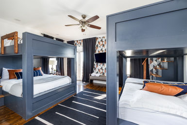 Cozy modern bunk room with navy built-in bunk beds, geometric accent wall, ceiling fan, hardwood floors, striped rug and wall-mounted TV.