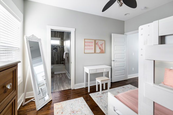Light-filled modern bedroom with white bunk bed and pink bedding, small white vanity and stool, leaning ornate full-length mirror, dark hardwood floors, gray walls, ceiling fan, and open door to a tiled ensuite bathroom.