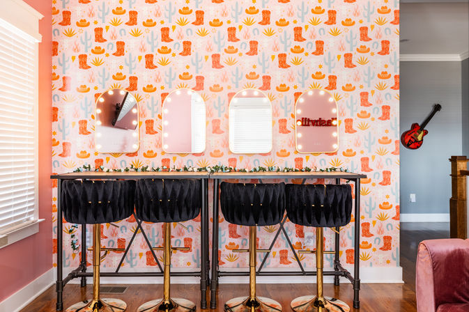 Playful pink home vanity with four lighted arched mirrors, western cowboy-boot and cactus wallpaper, a long high table with greenery, four black velvet bar stools with gold bases, and a red guitar on the adjacent wall.