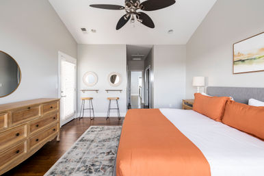 Bright modern master bedroom with king bed in orange bedding, gray upholstered headboard, natural wood dresser and nightstand, hardwood floors, patterned rug, ceiling fan, framed art, and two round mirrors above bar stools by a sunlit door.