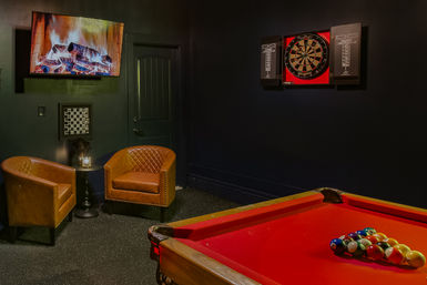 Cozy indoor game room with a red pool table racked for billiards, two brown leather club chairs by a small side table, a wall-mounted TV showing a fireplace, and a dartboard cabinet on a dark wall.