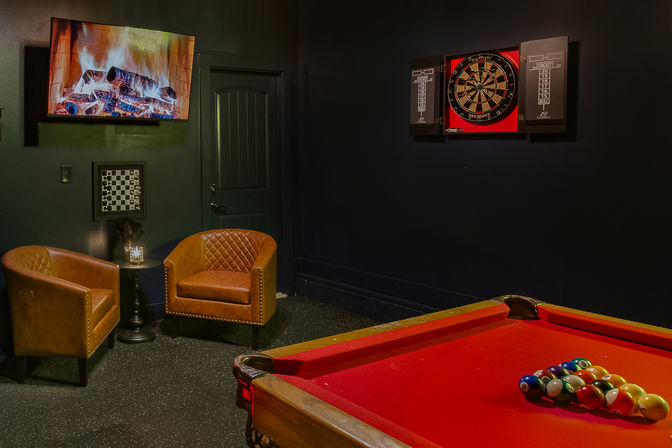 Cozy indoor game room with a red pool table racked for billiards, two brown leather club chairs by a small side table, a wall-mounted TV showing a fireplace, and a dartboard cabinet on a dark wall.