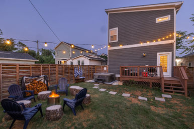 Cozy residential backyard at dusk with hanging string lights, a glowing fire pit surrounded by Adirondack chairs and log-stump seats, stacked firewood, a hot tub and wooden deck attached to a modern two-story gray house.