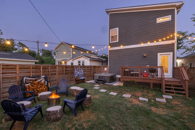 Cozy residential backyard at dusk with hanging string lights, a glowing fire pit surrounded by Adirondack chairs and log-stump seats, stacked firewood, a hot tub and wooden deck attached to a modern two-story gray house.