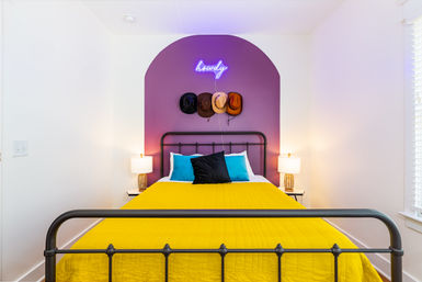 Playful modern bedroom with black iron bed, vibrant yellow quilt, teal and black pillows, purple arched accent wall topped by a neon “howdy” sign and four cowboy hats, flanked by bedside lamps.
