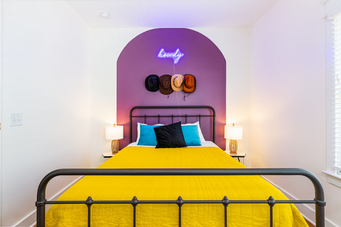 Playful modern bedroom with black iron bed, vibrant yellow quilt, teal and black pillows, purple arched accent wall topped by a neon “howdy” sign and four cowboy hats, flanked by bedside lamps.