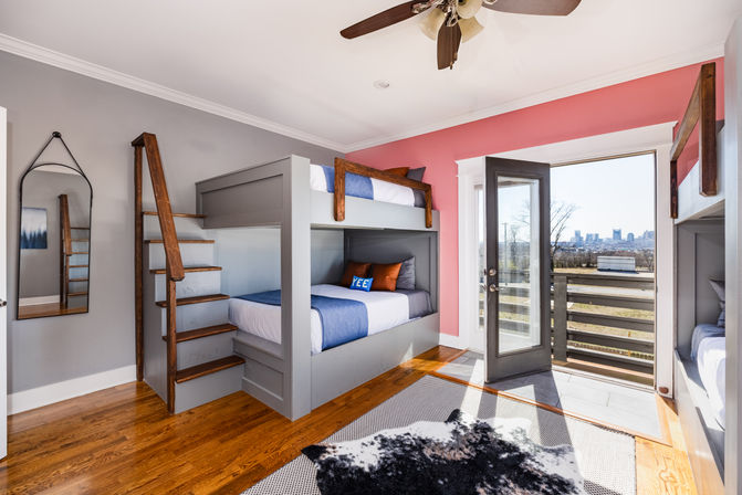 Bright bedroom with gray built-in double bunk beds and wooden ladder, blue-and-white bedding, coral accent wall, open glass door to balcony with distant city skyline, hardwood floors and ceiling fan.