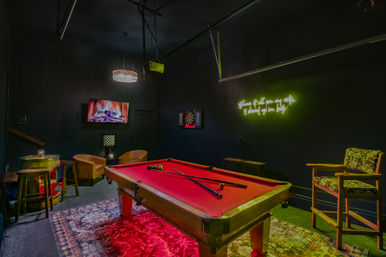 Red-felt pool table in a moody game room with neon green wall sign, wall-mounted TV displaying a fireplace, dartboard, leather lounge chairs, barrel-style high table, patterned rug and hanging chandelier.