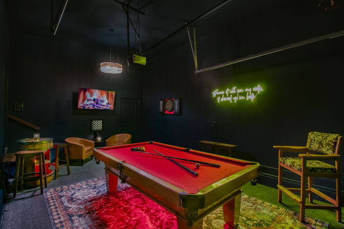 Red-felt pool table in a moody game room with neon green wall sign, wall-mounted TV displaying a fireplace, dartboard, leather lounge chairs, barrel-style high table, patterned rug and hanging chandelier.