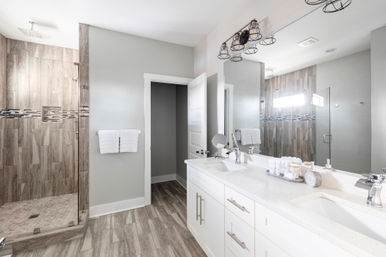 Bright, spa-like modern master bathroom with white double vanity and quartz countertop, large mirror, glass walk-in shower with wood-look tile and mosaic accent, towel rack and wood-look plank floor.